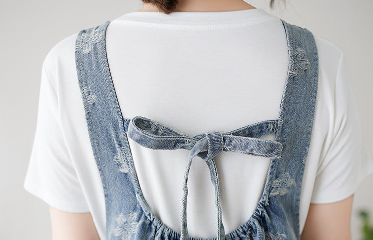 Denim pinafore dress with floral patterns worn over a white shirt on a neutral background.